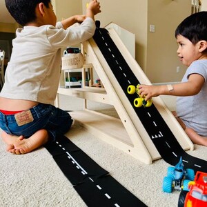 May include: Two children playing with a wooden toy garage and a black road mat. The road mat has white dashed lines. Yellow toy cars are on the road. The wooden garage has a sign that says "GARAGE".