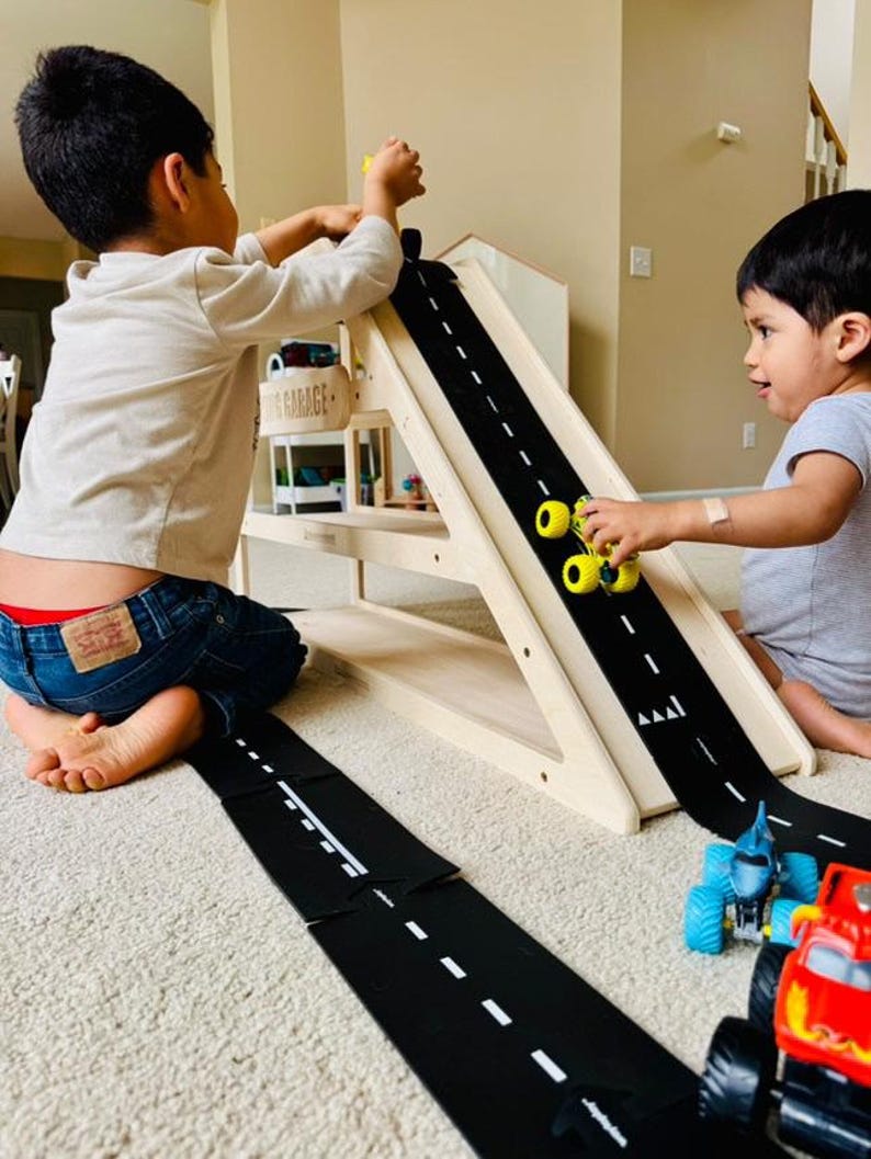 May include: Two children playing with a wooden toy garage and a black road mat. The road mat has white dashed lines. Yellow toy cars are on the road. The wooden garage has a sign that says "GARAGE".