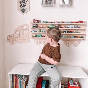 May include: A wooden toy truck shelf with a cutout of a truck on the left side. The shelf has four tiers and is filled with toy cars. A young child is sitting on a white bookshelf with a red speaker and toy cars on the shelves. The sign above the shelf says "Stay Rad".