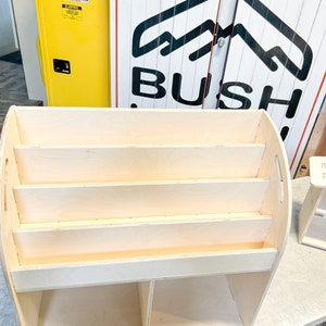 May include: A light-colored wooden bookshelf with four shelves and two lower compartments. The bookshelf has a curved top and sides with handles. The background includes a yellow cabinet with warning labels and a sign that says "BUSH."