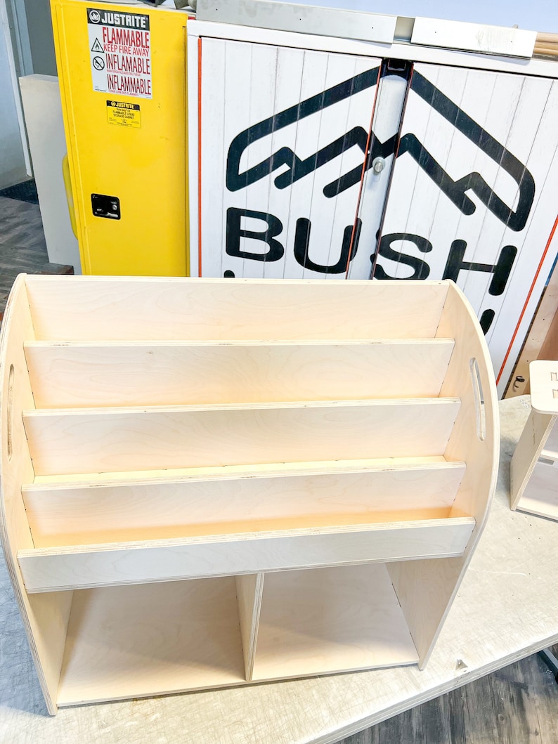 May include: A light-colored wooden bookshelf with four shelves and two lower compartments. The bookshelf has a curved top and sides with handles. The background includes a yellow cabinet with warning labels and a sign that says "BUSH."