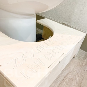 May include: A white wooden toilet seat riser with the words "ZXY I'M NOT A SPA" engraved on the top. The riser is sitting on a wooden floor.