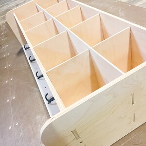May include: Unfinished wooden storage unit with multiple compartments. The unit has a light wood finish and is designed with a series of vertical dividers, creating individual storage spaces. A row of black hooks is attached to the side. The unit is ready for assembly.