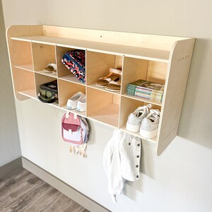 May include: A wooden wall-mounted organizer with multiple cubbies, holding shoes, a backpack, and books. A white jacket and a pink and white backpack hang below. The organizer is made of light-colored wood and is mounted on a neutral-colored wall.