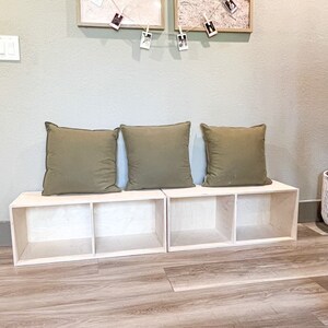 May include: A wooden bench with three storage compartments, each with a removable shelf. The bench is made of light wood and has a natural finish. Three green cushions are placed on top of the bench.