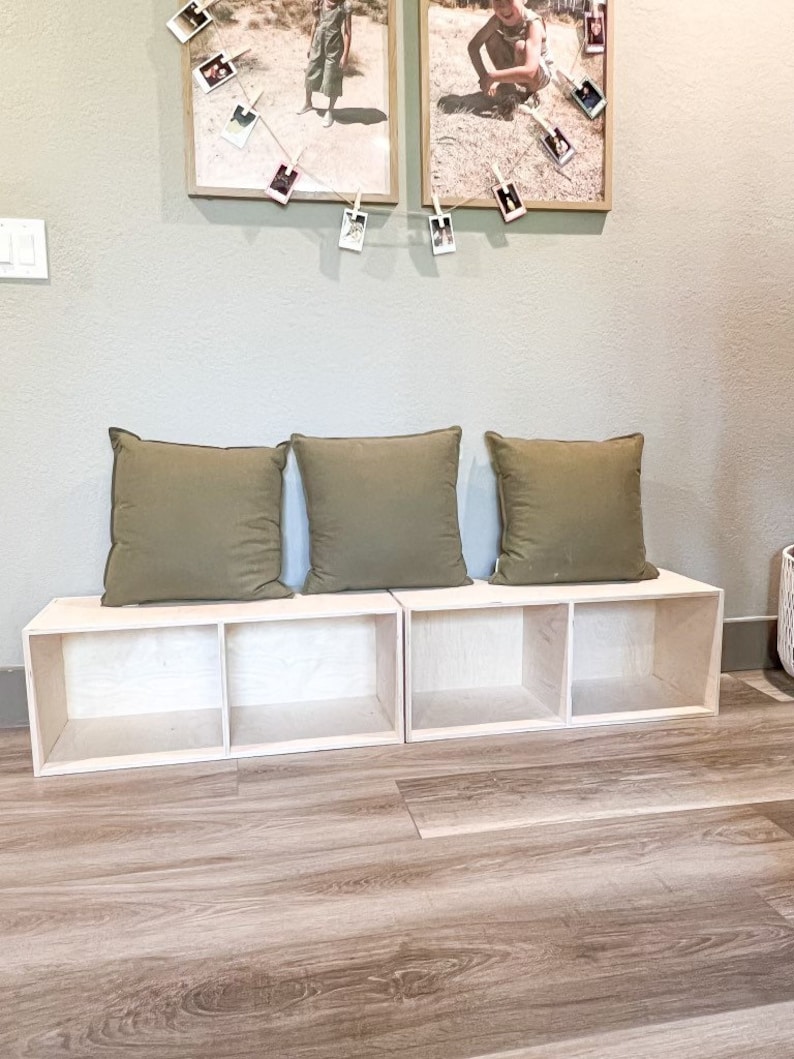 May include: A wooden bench with three storage compartments, each with a removable shelf. The bench is made of light wood and has a natural finish. Three green cushions are placed on top of the bench.