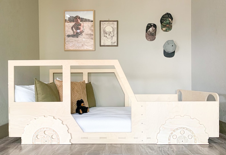 May include: A wooden bed frame shaped like a jeep, with a white mattress, green and brown pillows, and a black teddy bear. The bed is in a room with a light green wall and framed pictures.