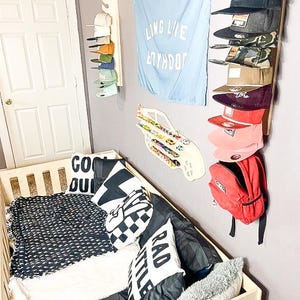 May include: A white crib with a black and white patterned blanket, a black and white checkered pillow, a white pillow with the text "RAD LITTLE" and a white pillow with the text "COOL DUDE". There is a red backpack hanging on the wall with a blue and white car toy on the wall above it. A blue flag with the text "LONG LIVE BOYHOOD" is hanging on the wall. There is a white shelf with a variety of hats on it.