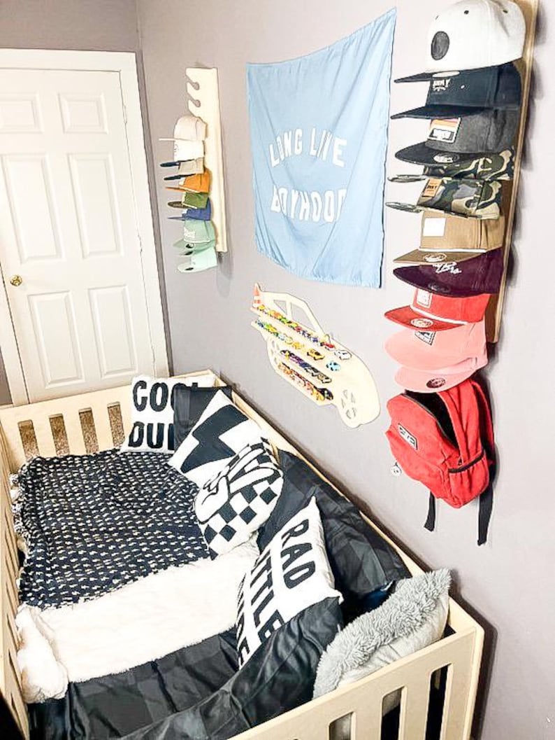 May include: A white crib with a black and white patterned blanket, a black and white checkered pillow, a white pillow with the text "RAD LITTLE" and a white pillow with the text "COOL DUDE". There is a red backpack hanging on the wall with a blue and white car toy on the wall above it. A blue flag with the text "LONG LIVE BOYHOOD" is hanging on the wall. There is a white shelf with a variety of hats on it.