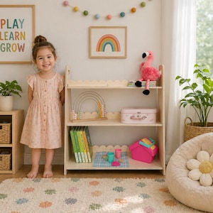 May include: A light wood bookshelf with three shelves, holding books, toys, and a pink suitcase. A young girl in a pink dress stands in front of the shelf. A rainbow art print and a felt ball garland decorate the wall.