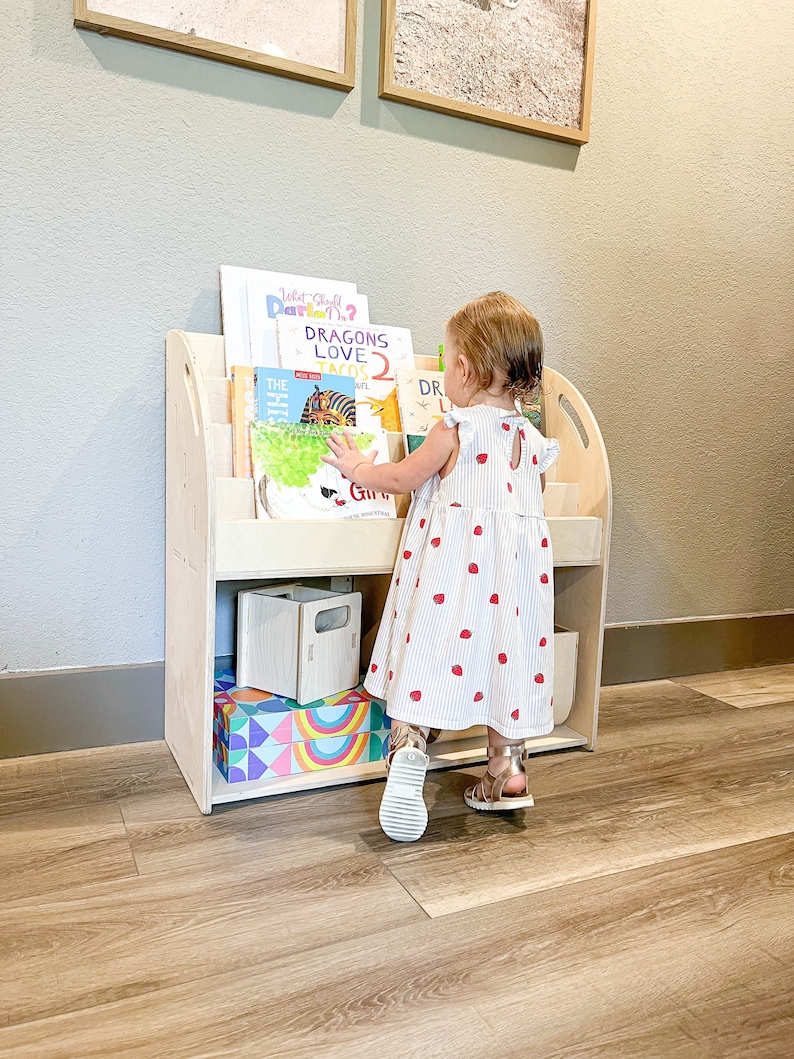 May include: A light-colored wooden bookshelf filled with children's books. A young child in a white dress with red strawberry prints is reaching for a book. The bookshelf has a curved top and several shelves.
