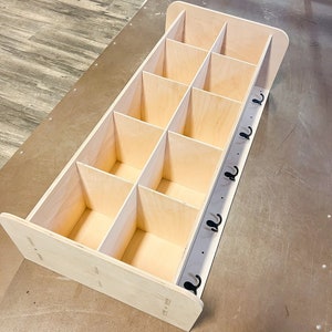 May include: A light-colored wooden storage unit with multiple cubbies and a row of black hooks. The unit is rectangular with rounded edges and is designed for organization and storage.