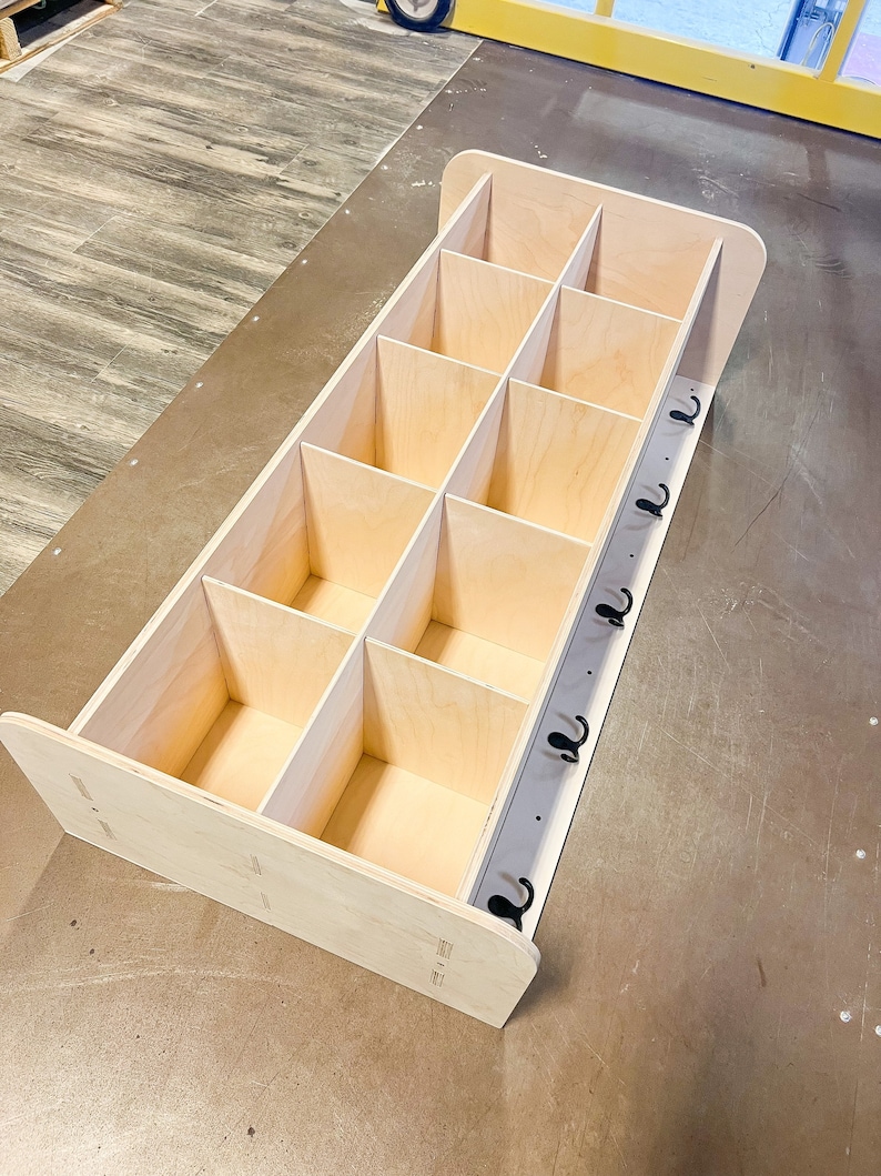 May include: A light-colored wooden storage unit with multiple cubbies and a row of black hooks. The unit is rectangular with rounded edges and is designed for organization and storage.