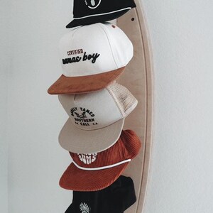 May include: A wooden surfboard-shaped hat rack with five baseball caps hanging on it. The caps are in various colors and styles, including black, white, brown, and beige. The caps have different logos and text, including "Certified mamas boy", "Wildly Tamed Southern CA", and "Gums".