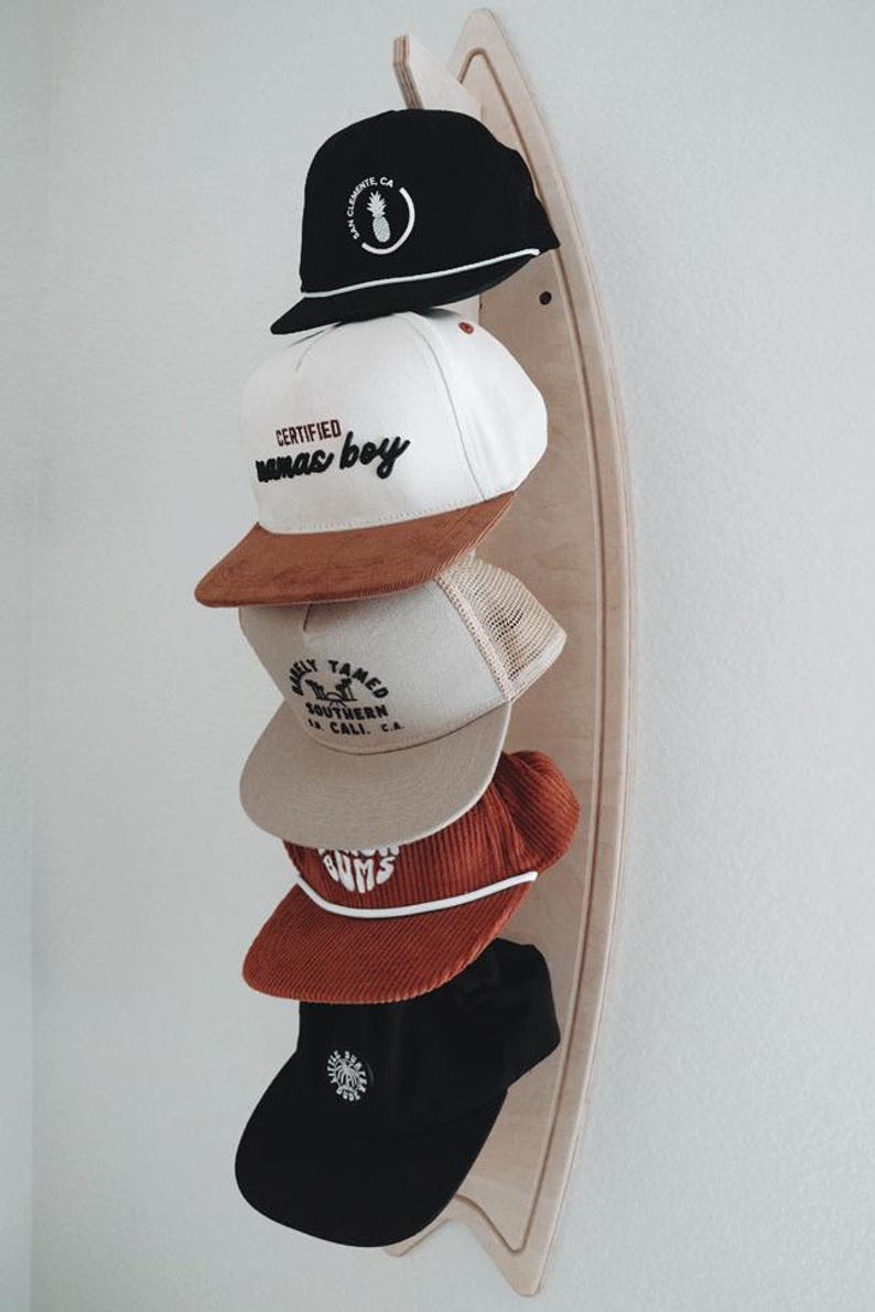 May include: A wooden surfboard-shaped hat rack with five baseball caps hanging on it. The caps are in various colors and styles, including black, white, brown, and beige. The caps have different logos and text, including "Certified mamas boy", "Wildly Tamed Southern CA", and "Gums".