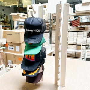 May include: A wooden hat rack with 10 slots for storing hats. Five hats are displayed on the rack, including a black hat with the text "Cowboy Hat" in white lettering, a green hat with a yellow and orange design, a black hat with the text "AZ" in rainbow lettering, a brown hat with a yellow leaf design, and a brown hat.