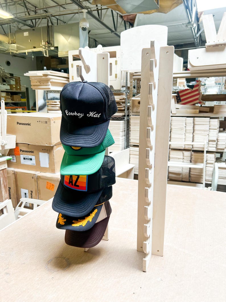 May include: A wooden hat rack with 10 slots for storing hats. Five hats are displayed on the rack, including a black hat with the text "Cowboy Hat" in white lettering, a green hat with a yellow and orange design, a black hat with the text "AZ" in rainbow lettering, a brown hat with a yellow leaf design, and a brown hat.