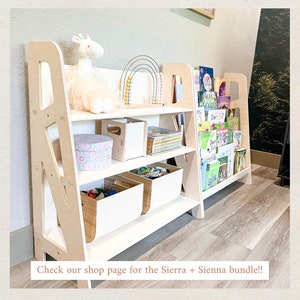 May include: A light-colored wooden bookshelf with multiple shelves. The shelves hold books, toys, and storage bins. A white llama plush toy and a rainbow abacus are on display. The bookshelf is designed for children's rooms.