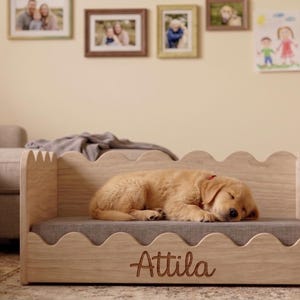 May include: A light-colored wooden pet bed with a scalloped top edge and a gray cushioned base. The front panel features the name Attila engraved in a cursive script. A golden-colored puppy is sleeping on the gray cushion inside the bed frame.