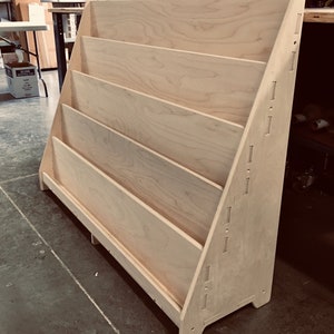 May include: A light wood bookshelf with five shelves. The shelves are angled to hold books or magazines. The bookshelf is made of plywood and has a natural finish.
