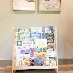 May include: A wooden bookshelf filled with children's books. The bookshelf is light-colored and has a slanted design. The books have colorful covers with titles like "Home in the Woods", "Princess", and "Treasure Island". Two framed photos hang above the bookshelf.