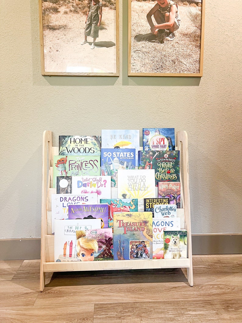 May include: A wooden bookshelf filled with children's books. The bookshelf is light-colored and has a slanted design. The books have colorful covers with titles like "Home in the Woods", "Princess", and "Treasure Island". Two framed photos hang above the bookshelf.