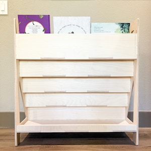 May include: A light-colored wooden bookshelf with a tiered design. The bookshelf has a rectangular base and angled sides. Several books are visible on the top shelf. The text on the books is visible. The bookshelf is made of light-colored wood.