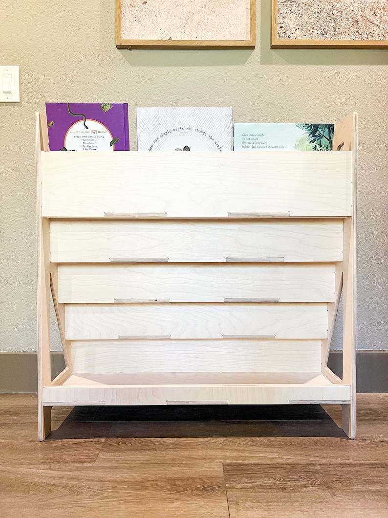 May include: A light-colored wooden bookshelf with a tiered design. The bookshelf has a rectangular base and angled sides. Several books are visible on the top shelf. The text on the books is visible. The bookshelf is made of light-colored wood.