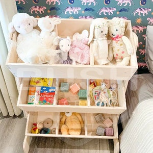 May include: A white wooden toy shelf with four compartments. The top compartment has stuffed animals, including a white lamb, a white bunny, a pink bunny, and a grey mouse. The second compartment has books, blocks, and a giraffe toy. The third compartment has a brown teddy bear and wooden blocks. The bottom compartment has wooden toys.