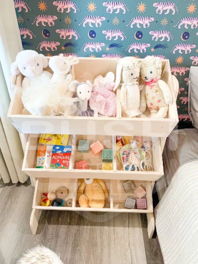 May include: A white wooden toy shelf with four compartments. The top compartment has stuffed animals, including a white lamb, a white bunny, a pink bunny, and a grey mouse. The second compartment has books, blocks, and a giraffe toy. The third compartment has a brown teddy bear and wooden blocks. The bottom compartment has wooden toys.