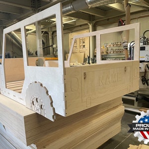May include: A wooden cutout of a jeep bed frame with the word "NAME" carved into the back. The frame is made of plywood and is sitting on a stack of plywood sheets. The frame is in a workshop setting.
