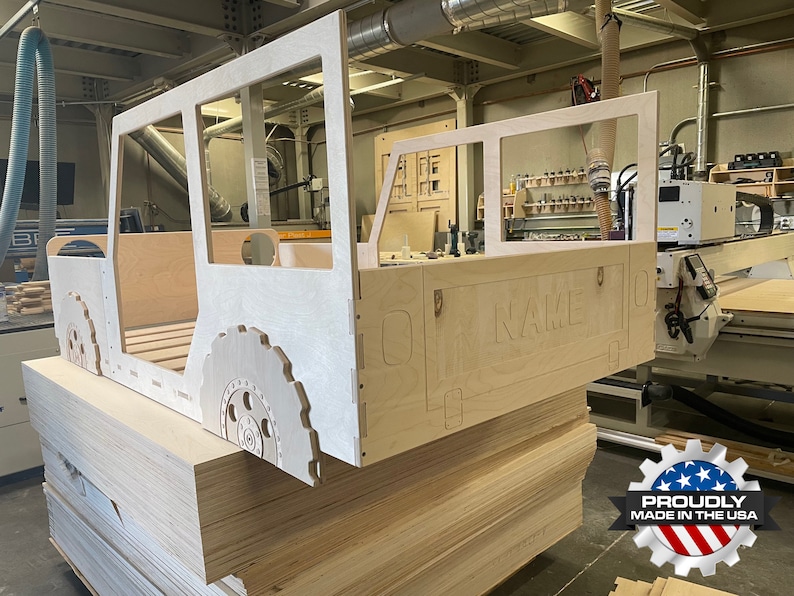 May include: A wooden cutout of a jeep bed frame with the word "NAME" carved into the back. The frame is made of plywood and is sitting on a stack of plywood sheets. The frame is in a workshop setting.