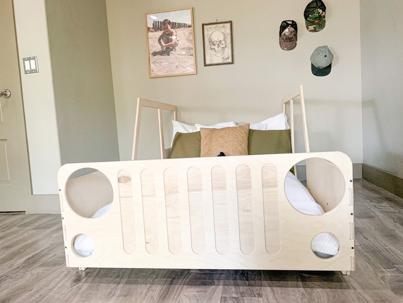 May include: A wooden bed frame shaped like a jeep, with a white mattress and pillows. The bed is in a room with a light brown wood floor and white walls.
