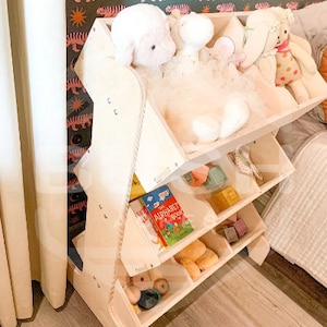 May include: A wooden toy shelf with multiple compartments, featuring a book titled "The Alphabet Book" and stuffed animals. The shelf is made of light wood and has a natural finish.
