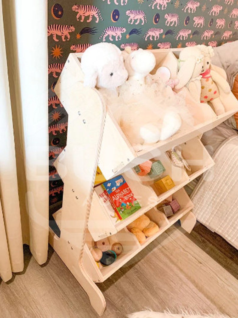 May include: A wooden toy shelf with multiple compartments, featuring a book titled "The Alphabet Book" and stuffed animals. The shelf is made of light wood and has a natural finish.