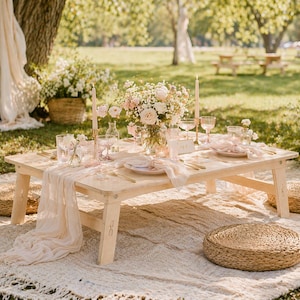 May include: A low, light-colored wooden picnic table set for a gathering. The table is adorned with a floral arrangement, candles, and place settings. Woven floor cushions and a cream-colored rug complete the outdoor scene.
