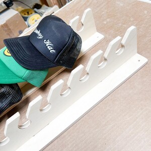 May include: A white wooden hat rack with six slots for storing hats. A black baseball cap with the words "Weekday Hat" embroidered on it is hanging on the rack.