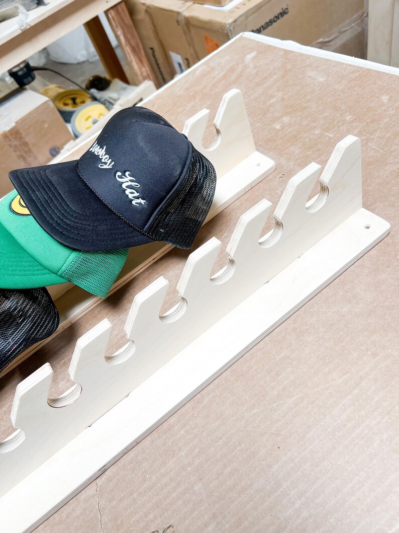 May include: A white wooden hat rack with six slots for storing hats. A black baseball cap with the words "Weekday Hat" embroidered on it is hanging on the rack.
