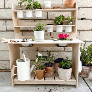 May include: A wooden play kitchen with a shelf for gardening. The shelf has several small white pots with plants. The kitchen has two stainless steel bowls and a white watering can. There are several plants in pots on the bottom shelf.