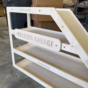 May include: A wooden toy parking garage with three shelves and a ramp. The words "PARKING GARAGE" are carved into a horizontal sign. The structure is made of light-colored wood and has a simple, modern design.