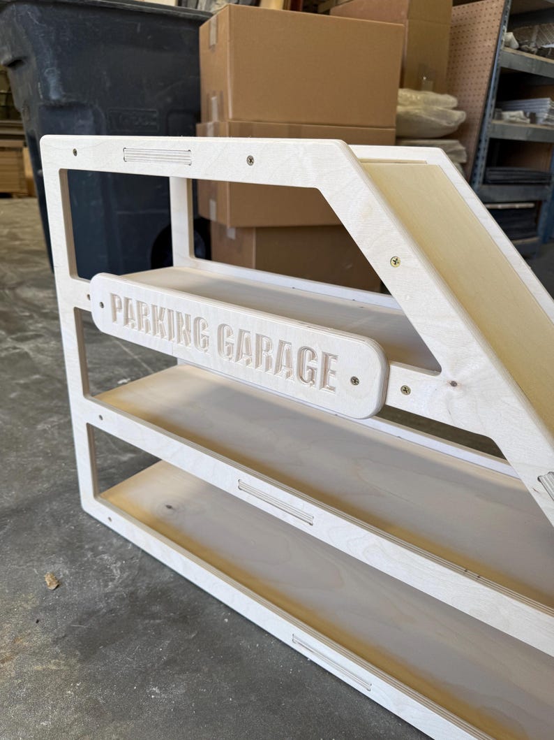 May include: A wooden toy parking garage with three shelves and a ramp. The words "PARKING GARAGE" are carved into a horizontal sign. The structure is made of light-colored wood and has a simple, modern design.