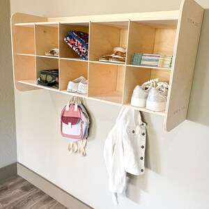 May include: A light-colored wooden wall-mounted storage unit with multiple cubbies. The unit holds shoes, books, and a backpack. A white denim jacket hangs below. The unit is mounted on a light gray wall.