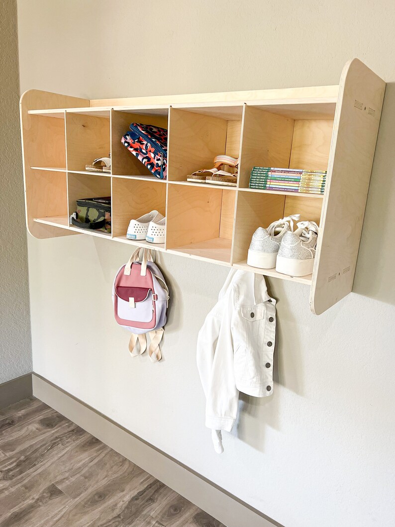 May include: A light-colored wooden wall-mounted storage unit with multiple cubbies. The unit holds shoes, books, and a backpack. A white denim jacket hangs below. The unit is mounted on a light gray wall.