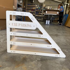 May include: A wooden, triangular-shaped toy with four shelves. The sign on the front reads "CAR PARKING". The toy is made of light-colored wood and is designed for children's play.