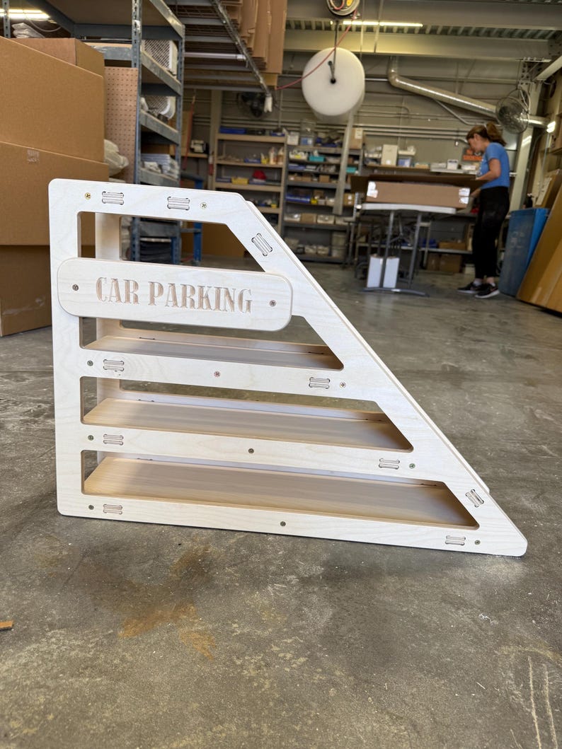 May include: A wooden, triangular-shaped toy with four shelves. The sign on the front reads "CAR PARKING". The toy is made of light-colored wood and is designed for children's play.