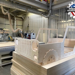 May include: A wooden bed frame shaped like a Jeep, with a slatted headboard and a detailed tyre design on the side. The bed frame is unfinished and is sitting on a stack of plywood sheets in a workshop setting. A sign in the background reads "Proudly Made in the USA."