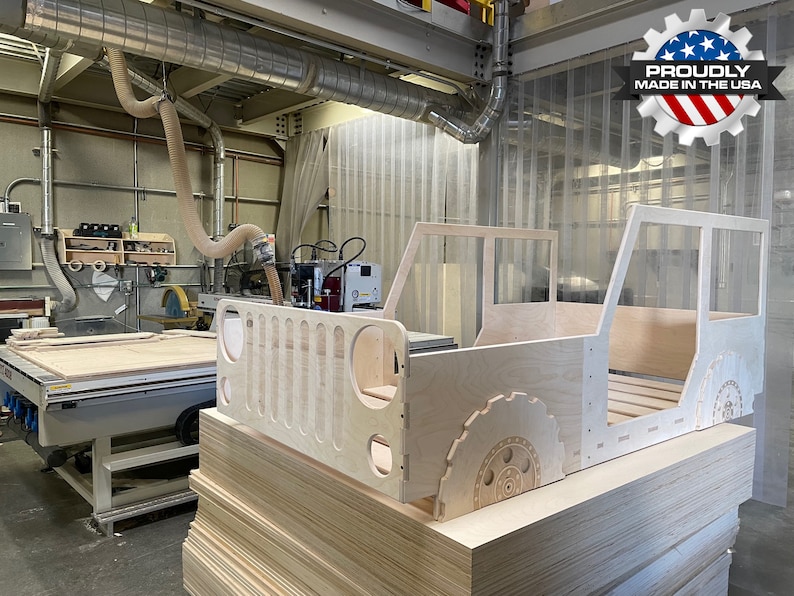 May include: A wooden bed frame shaped like a Jeep, with a slatted headboard and a detailed tyre design on the side. The bed frame is unfinished and is sitting on a stack of plywood sheets in a workshop setting. A sign in the background reads "Proudly Made in the USA."