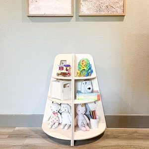 May include: A wooden, triangular-shaped bookshelf with five shelves. The shelves are filled with toys, books, and art supplies. The bookshelf is standing on a wooden floor.