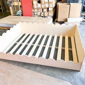 May include: A light-colored wooden bed frame with a scalloped edge design. The bed frame has a slatted base and is constructed from unfinished wood. The frame is rectangular and appears to be for a twin or full-size bed.