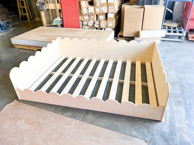 May include: A light-colored wooden bed frame with a scalloped edge design. The bed frame has a slatted base and is constructed from unfinished wood. The frame is rectangular and appears to be for a twin or full-size bed.